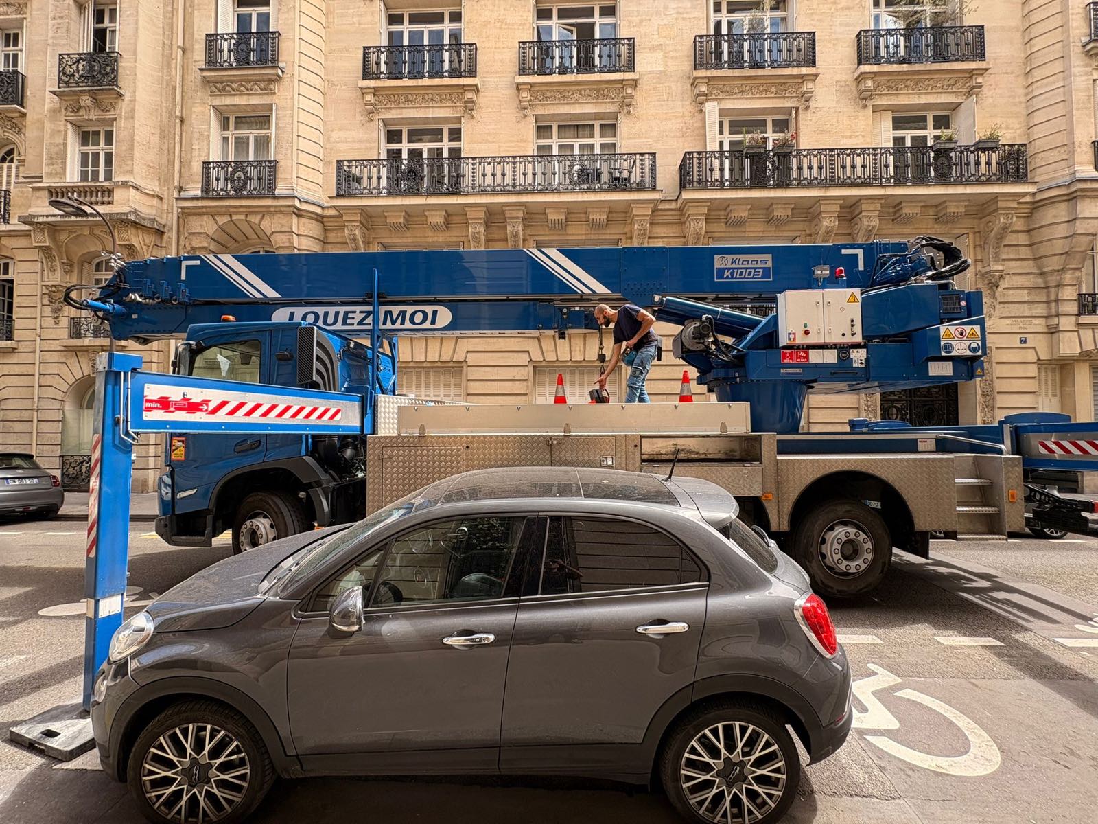 Grue sur camion Klaas K1003 d'AÉRO LEVAGE repliée devant une façade haussmannienne, opérateur préparant l'élingage sur le plateau