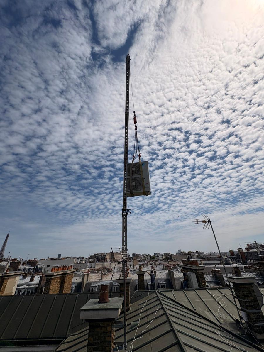 Groupe de climatisation élingué en bout de câble au-dessus des toits de zinc parisiens, deux techniciens en réception sur la toiture, Paris 16e