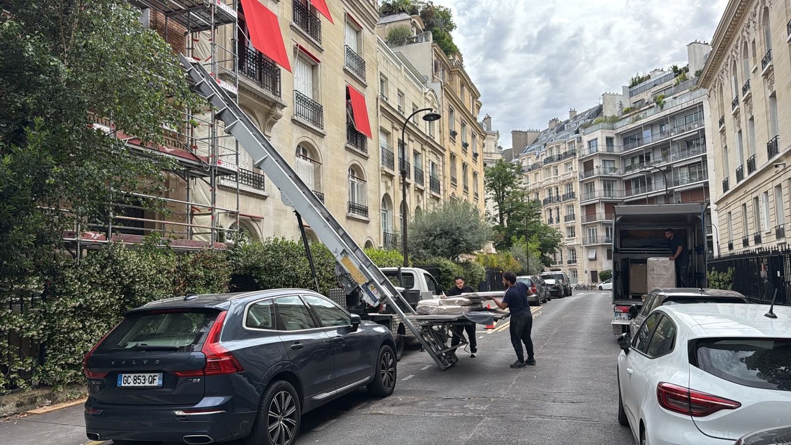 Monte-meuble déployé le long d'une façade haussmannienne sous échafaudage, rue du 16e arrondissement de Paris, deux opérateurs chargeant des éléments filmés sur le plateau